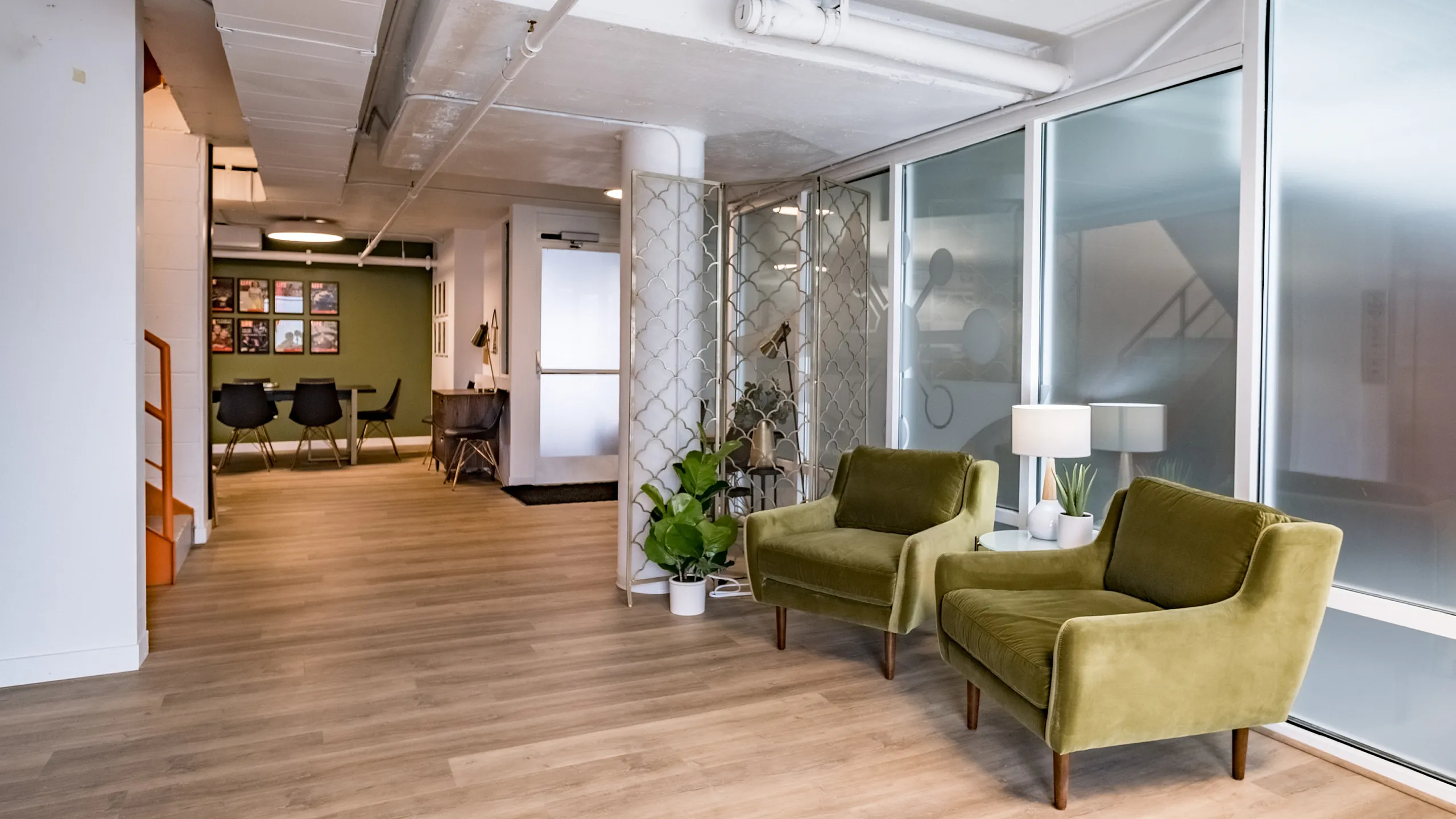 Spacious hallway with wooden flooring, featuring two olive green chairs and a white lamp by a large frosted window. Green wall and chairs in distance.