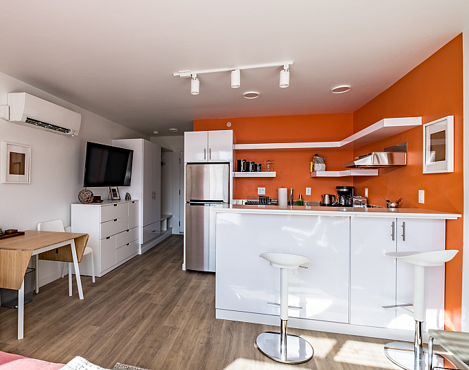 Modern studio apartment with an open layout, featuring a bright orange accent wall in the kitchen, sleek white cabinets, a compact dining area, and cozy furnishings.