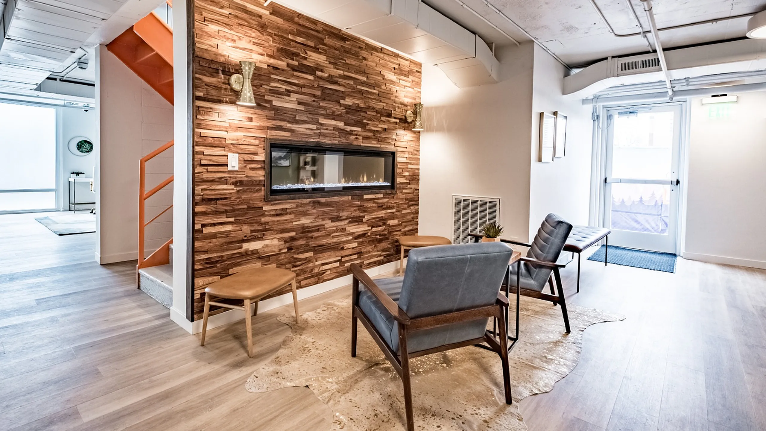 Modern lounge area with wooden accent wall showcasing a fireplace. Features two blue chairs and wooden stools on a light wood floor with cozy ambiance.