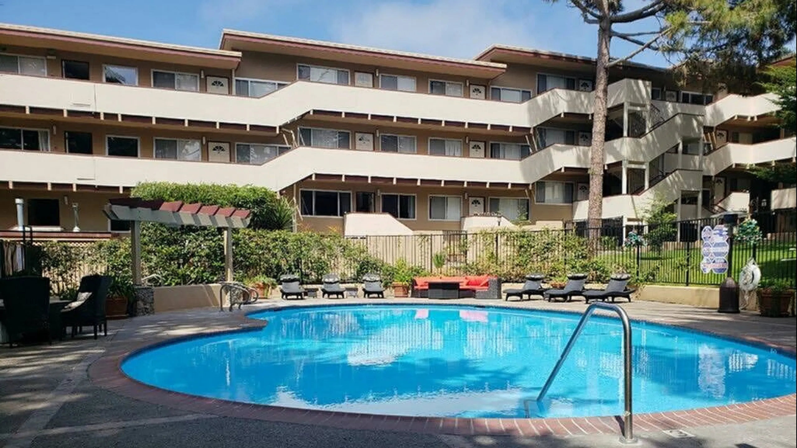 An exterior view of a sunny swimming pool area surrounded by a brick deck, lounge chairs, and a multi-story apartment complex with lush trees in the background.