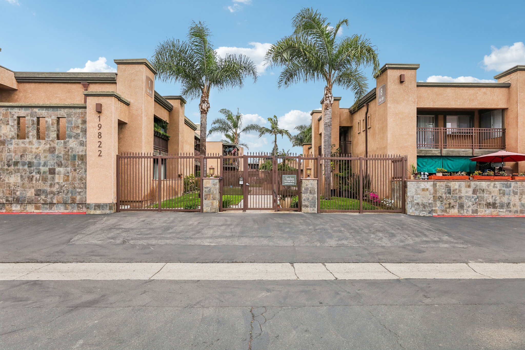 Charming apartment complex entrance with stone features, gated access, and lush palm trees under a clear blue sky.