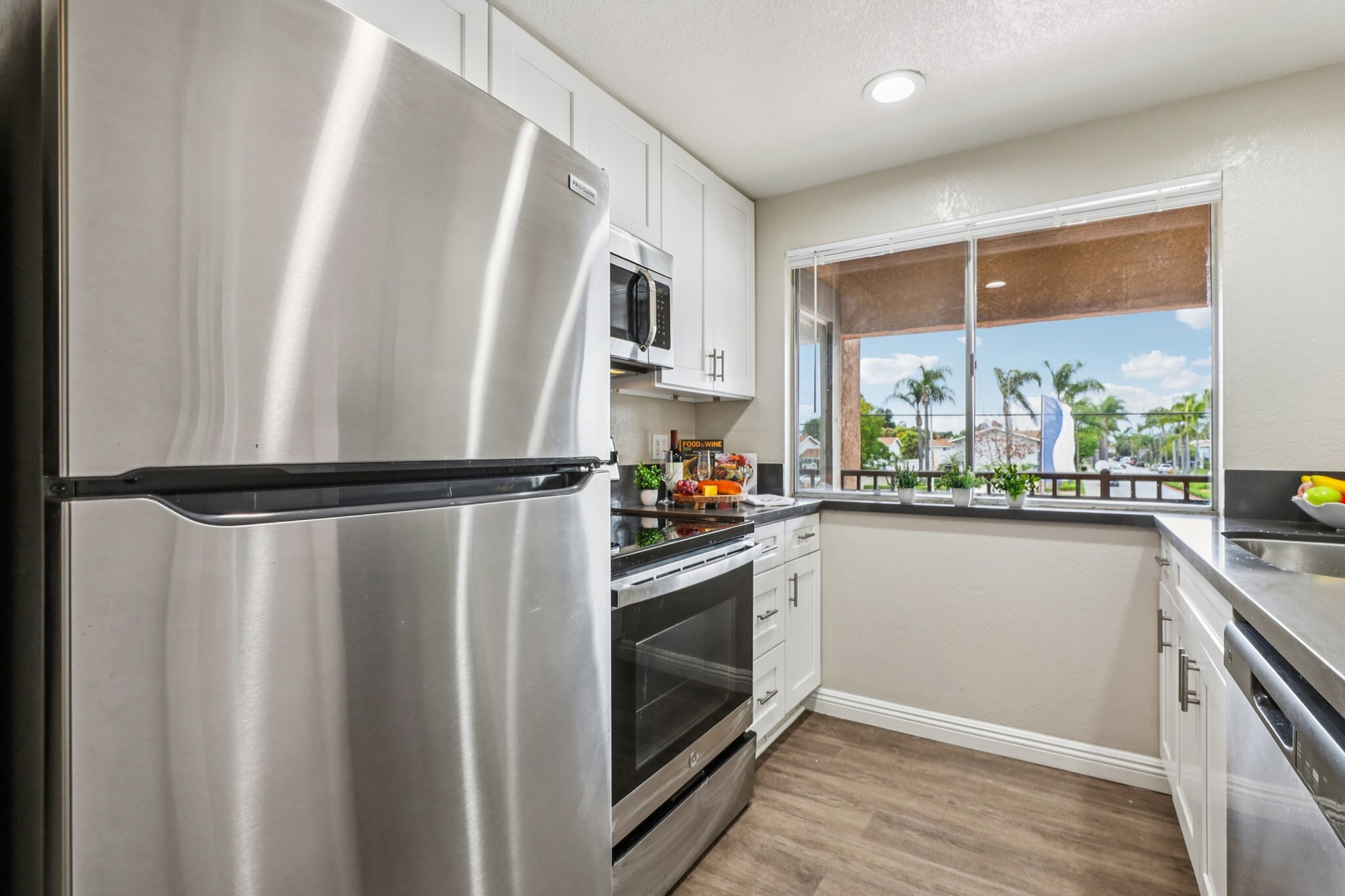 Modern kitchen with sleek stainless steel appliances, white cabinetry, and a bright window view of palm trees.