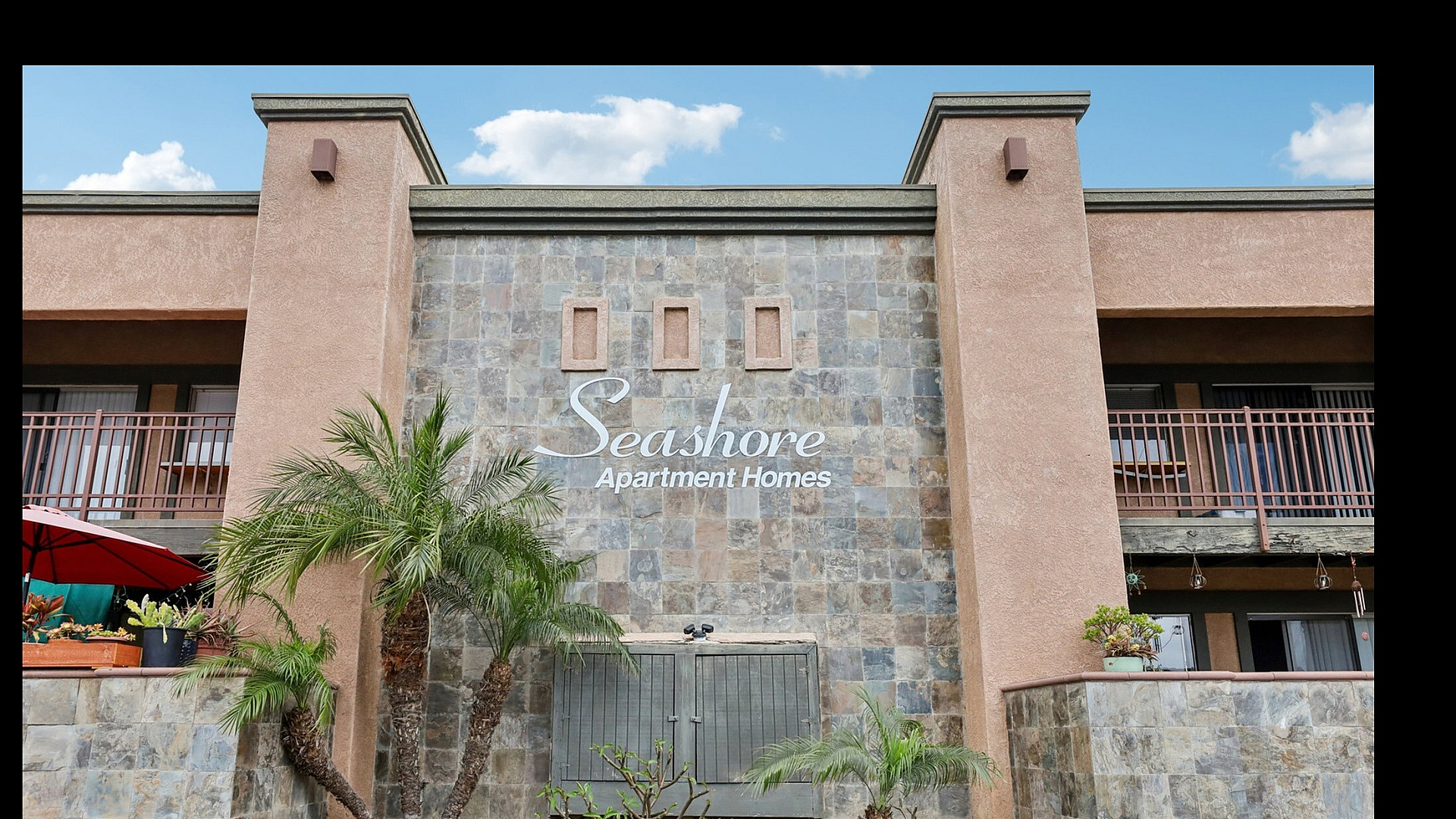 Charming facade of Seashore Apartment Homes featuring stone accents, lush palm trees, and cozy balconies offering a welcoming atmosphere.