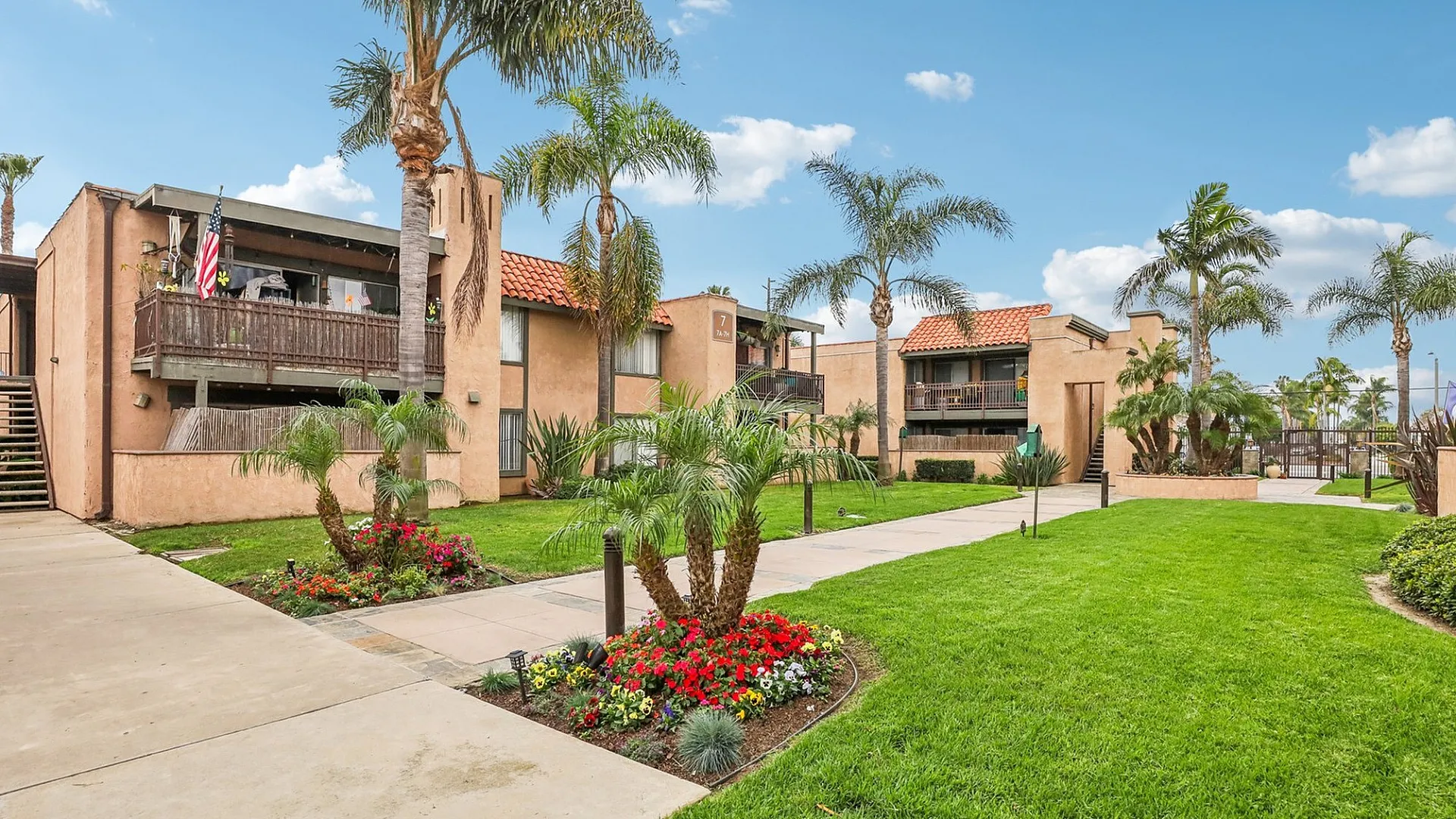Stunning apartment complex surrounded by lush green lawns, vibrant flower beds, and tall palm trees, featuring a warm exterior with charming wooden balconies.