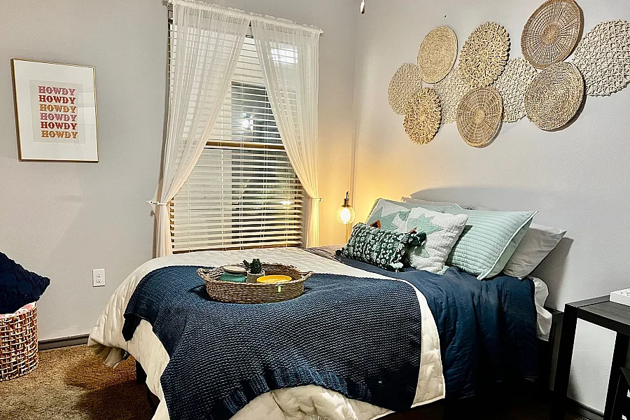 Cozy bedroom featuring a plush bed with blue and white bedding, stylish woven wall decor, and warm ambient lighting.