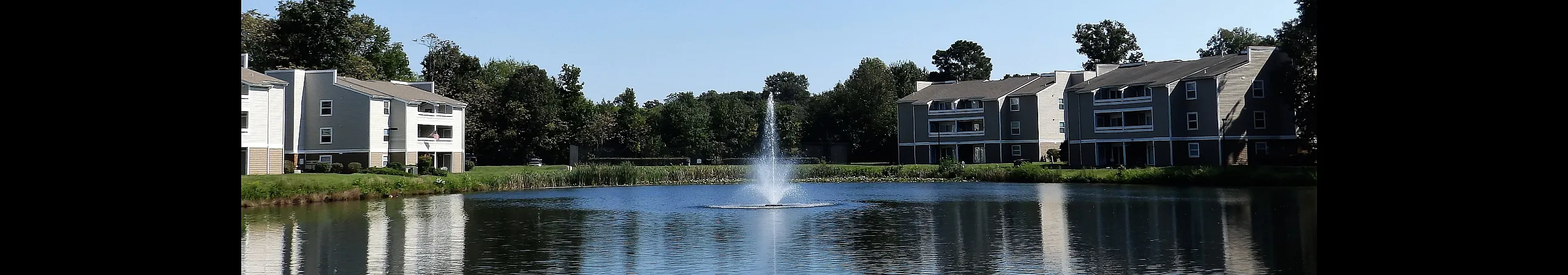 Serene lakeside apartment community with elegant buildings, lush greenery, and a central fountain enhancing the tranquil atmosphere.