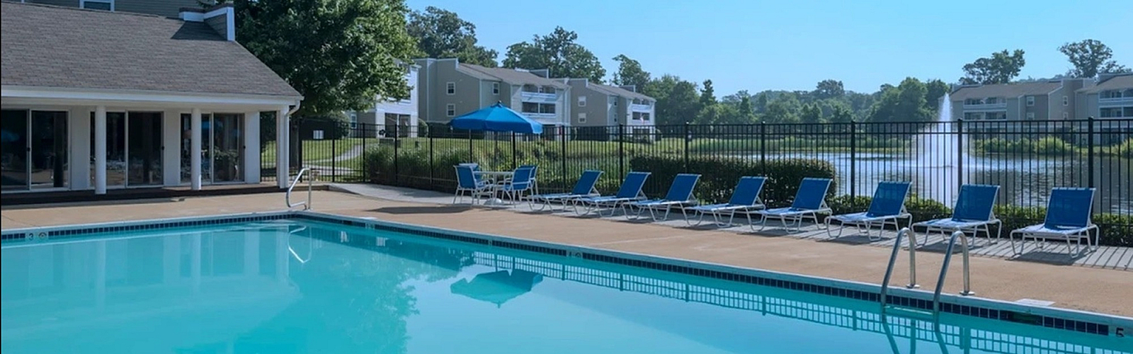 Inviting apartment complex with a sparkling pool, modern lounge chairs, and scenic pond view, perfect for relaxation and leisure.