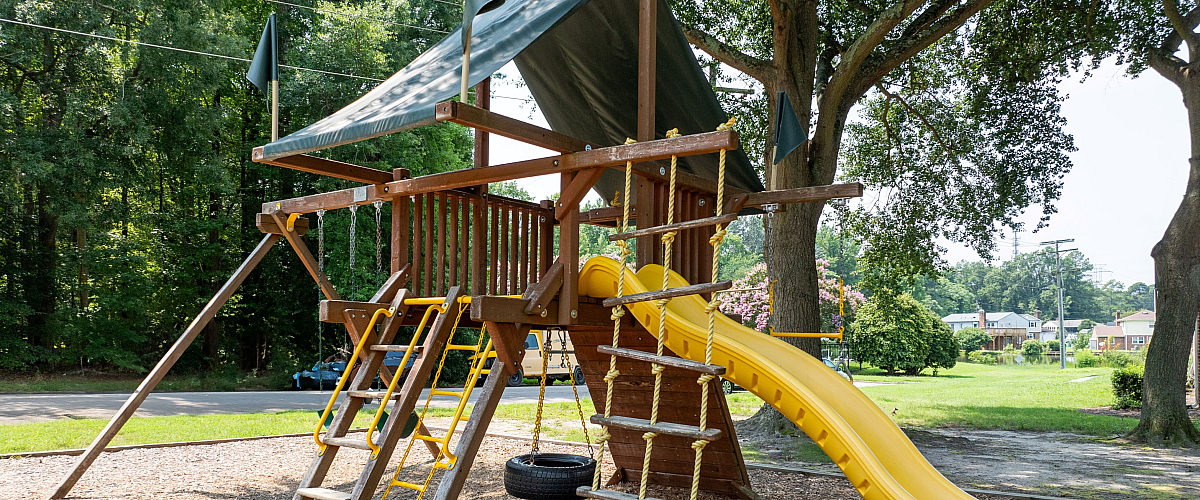 Charming playground with a wooden play structure featuring a vibrant yellow slide, rope ladder, and shaded canopy, surrounded by lush greenery.