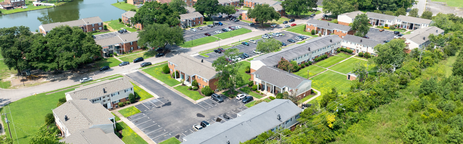 Aerial view of a charming apartment community with lush greenery, spacious parking, and a serene lake backdrop.