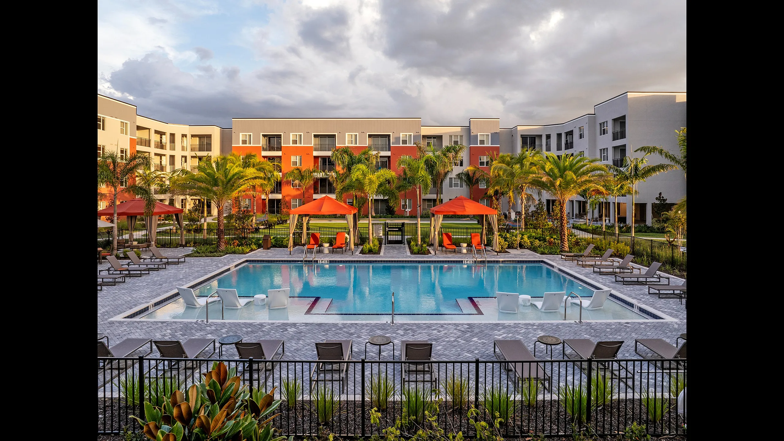 Luxurious apartment complex featuring a pristine swimming pool with red cabana loungers and lush palm trees.