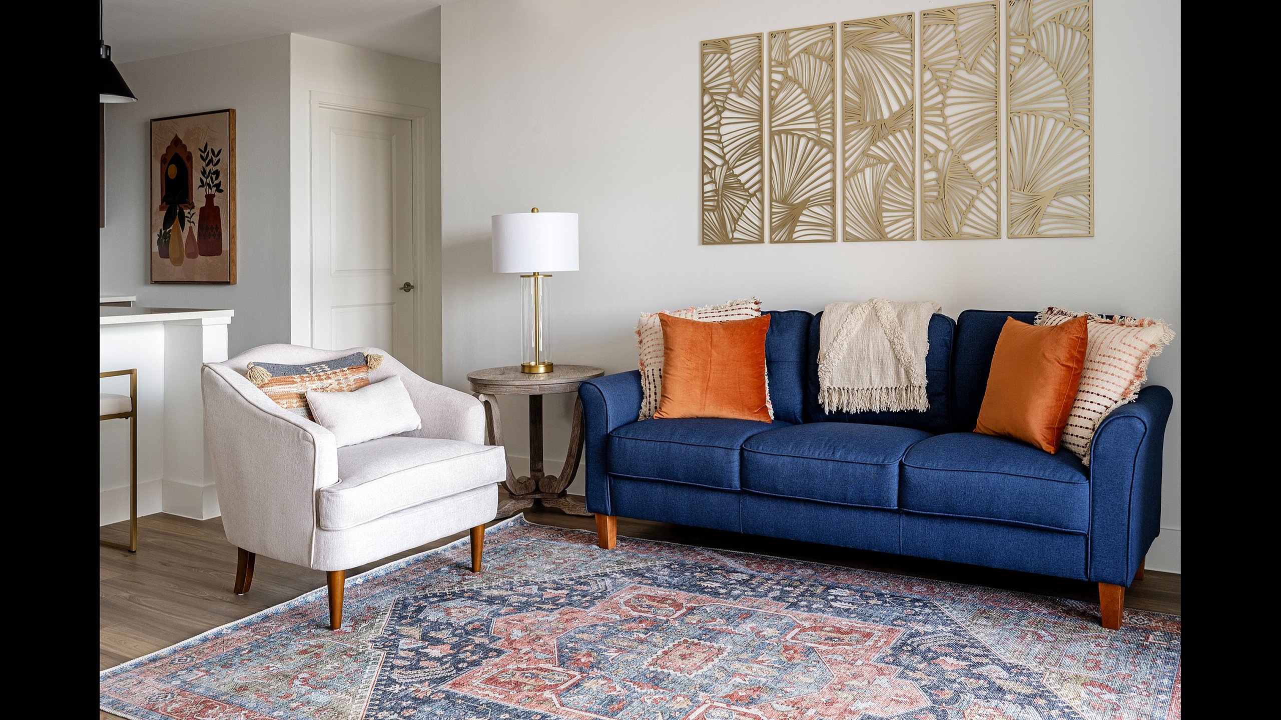 Stylish living room with a blue couch, cream armchair, and elegant wall art, featuring warm-toned pillows and a patterned rug.