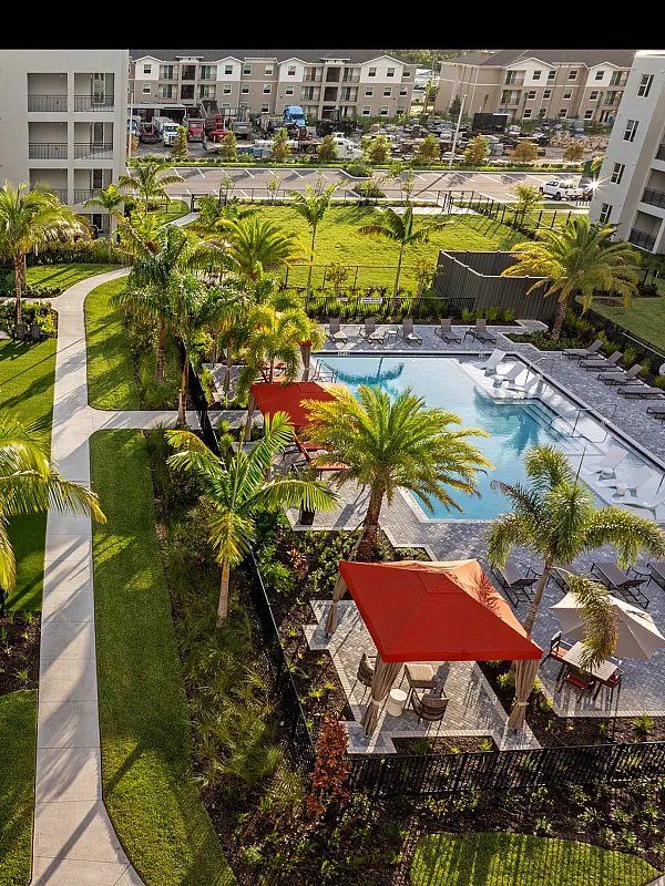 Aerial view of a modern apartment complex featuring lush gardens, a pathway, and a sparkling swimming pool with red cabanas.