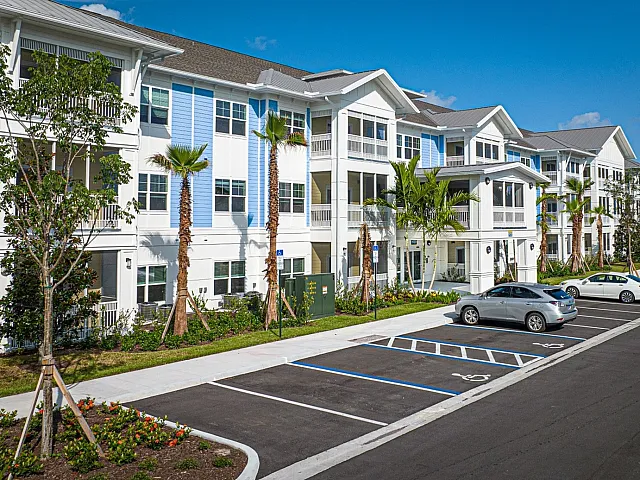 Charming modern apartment building with bright, fresh facade, surrounded by lush landscaping and ample parking, under a clear blue sky.