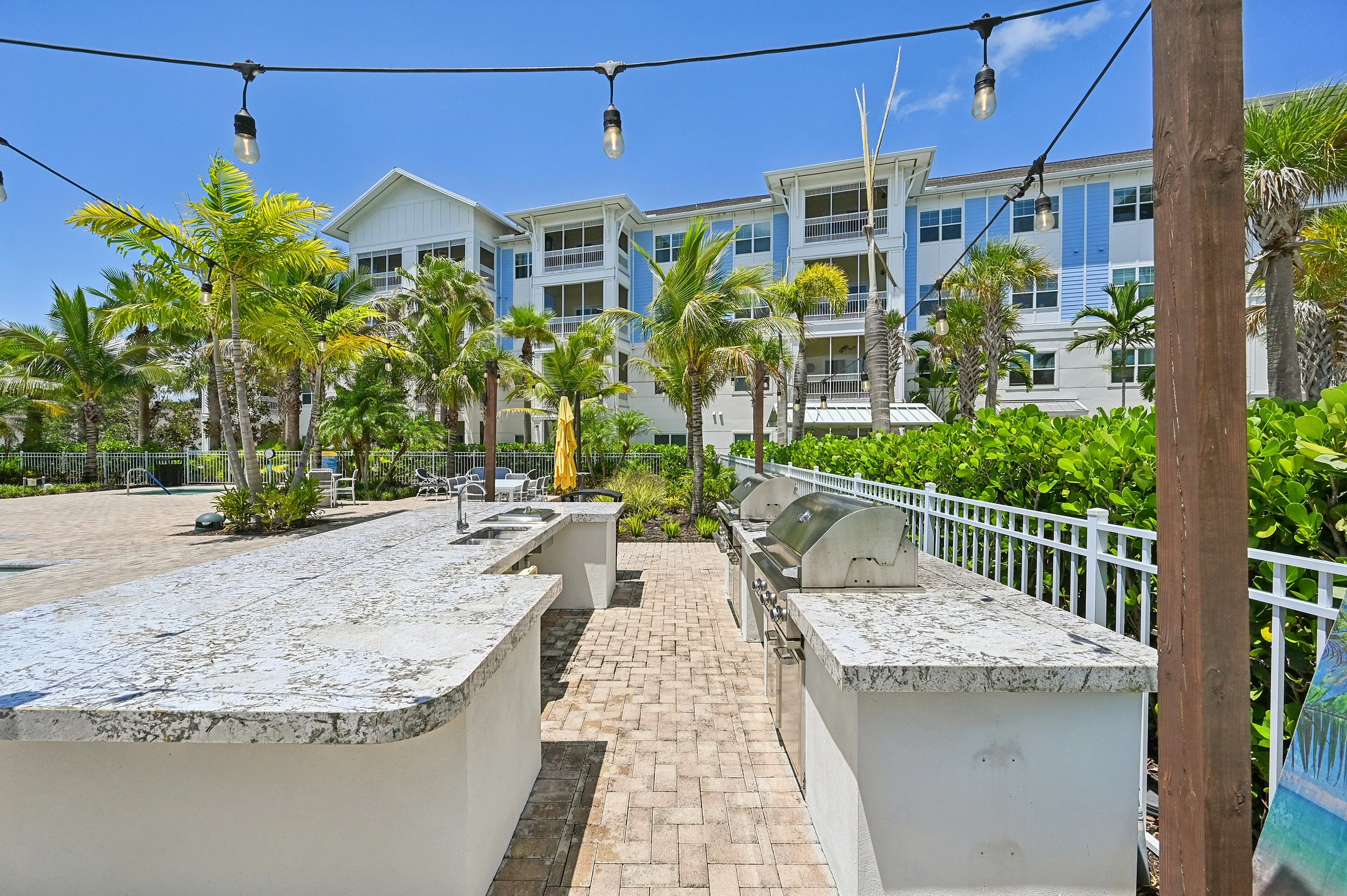 Charming outdoor cooking area with sleek granite countertops, modern grills, and lush palm trees in a resort-style apartment setting under a vibrant blue sky.