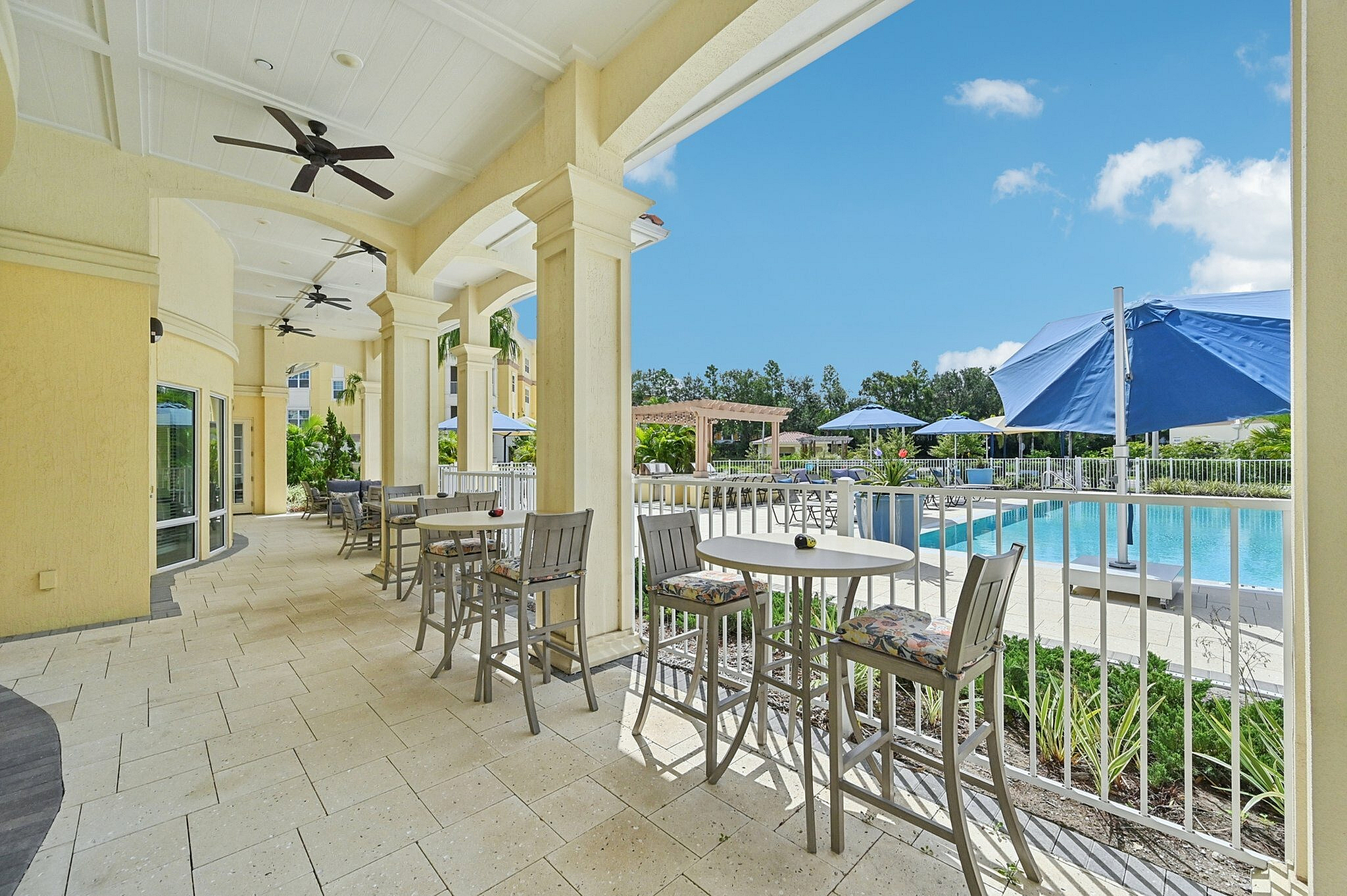 Luxurious poolside patio with elegant seating, ceiling fans, and vibrant blue umbrellas set against a clear sky, perfect for relaxing and enjoying the sunshine.