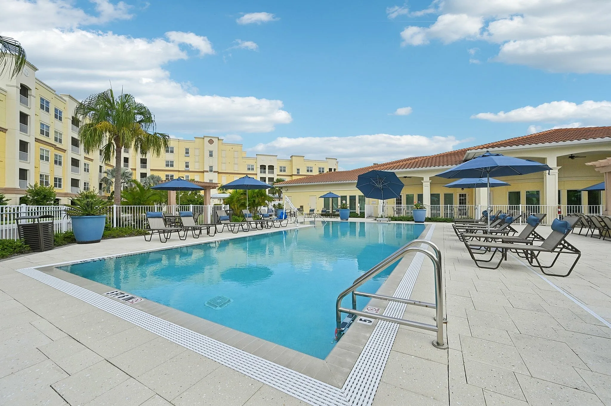 Luxurious apartment complex showcasing a sparkling pool with elegant lounge chairs and vibrant blue umbrellas amid lush greenery.