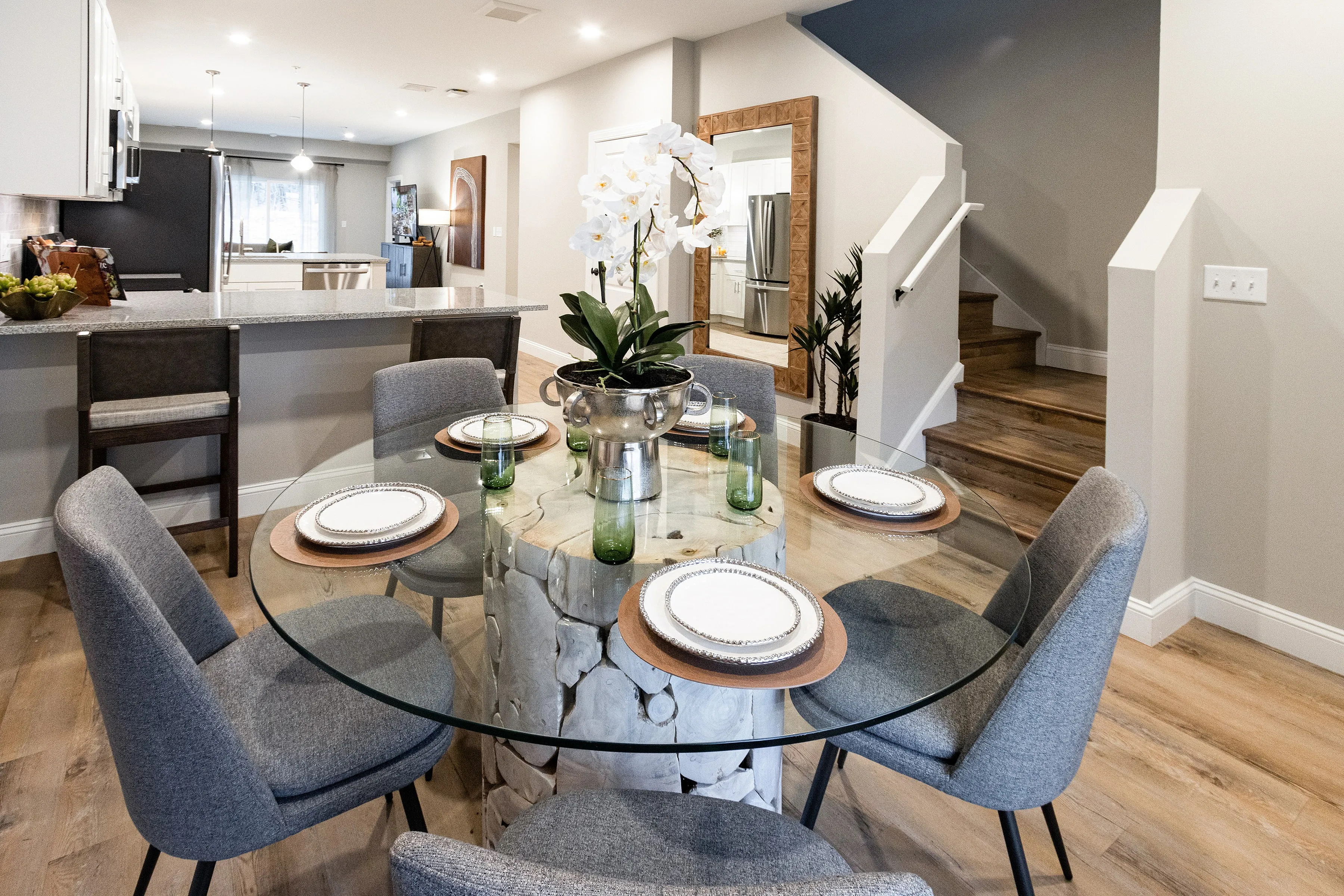 Contemporary dining area with elegant glass-top table, stylish gray chairs, and a view into a modern open-plan kitchen with sleek bar seating.