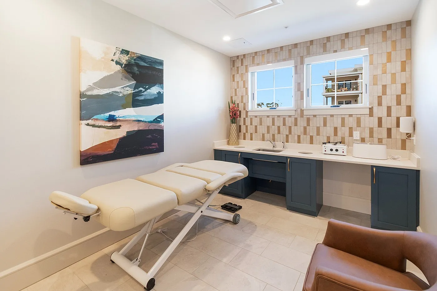 Elegant and serene spa room featuring a plush massage table, modern tiled backsplash, and artistic wall decor. Natural light flows through large windows, creating a peaceful ambiance.