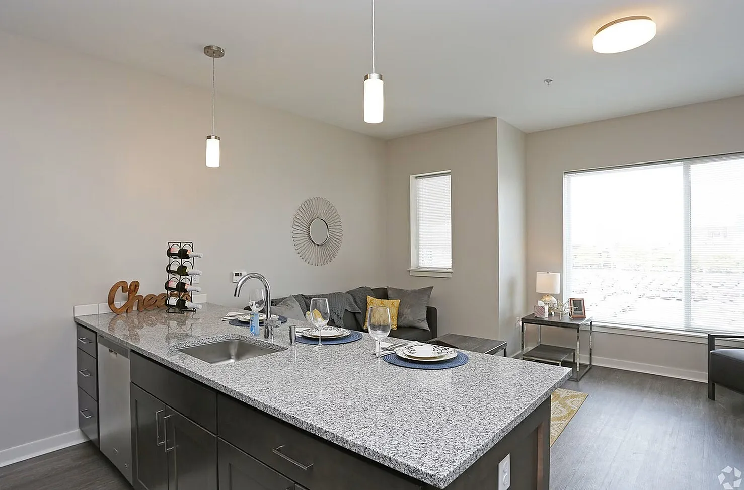 Modern apartment with open-concept kitchen and living room, featuring a sleek granite countertop, stylish pendant lighting, and bright natural light from large windows.