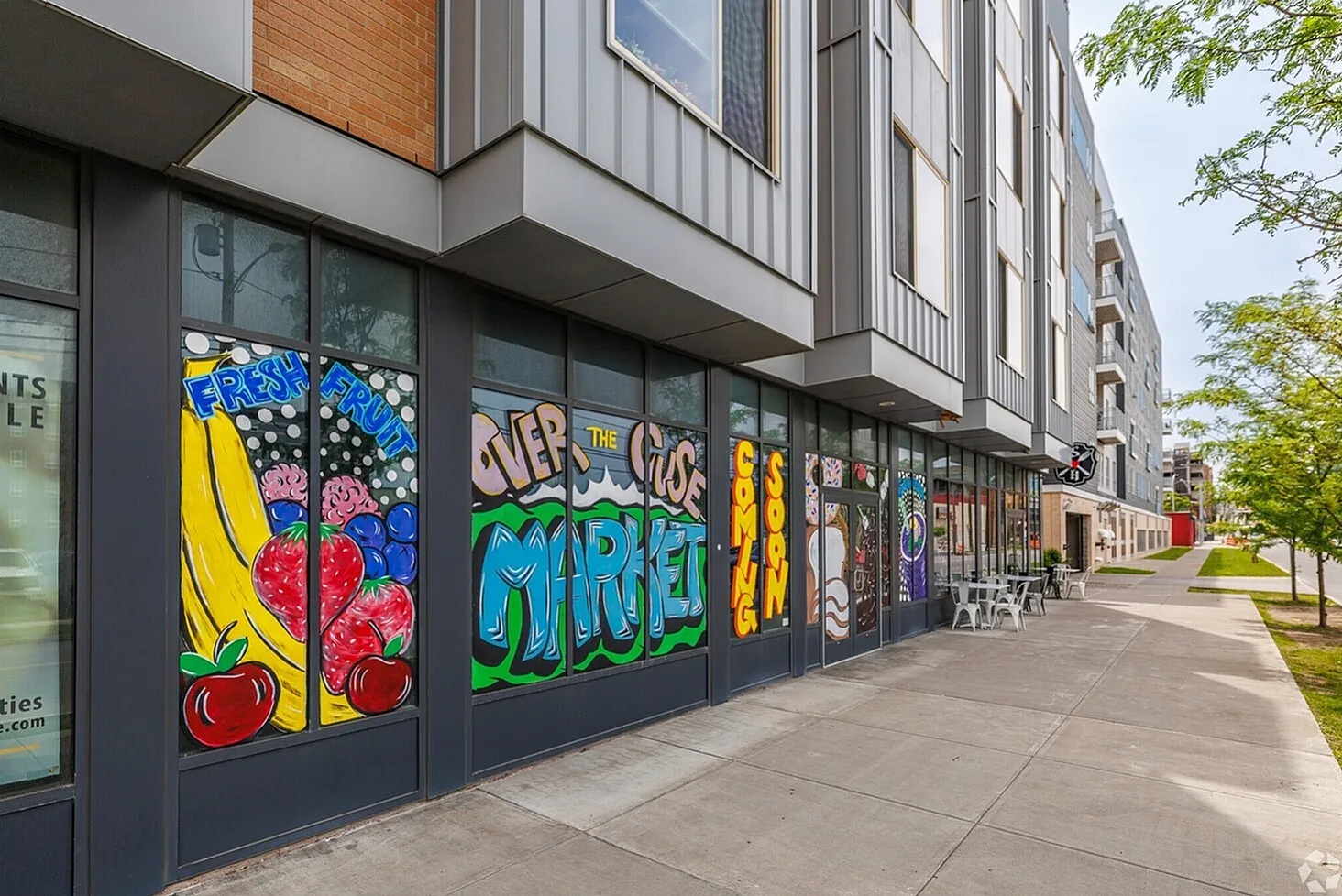 Vibrant urban setting with artistic murals on storefront windows, featuring fresh fruits and bold typography, promising an upcoming market in a contemporary apartment building.