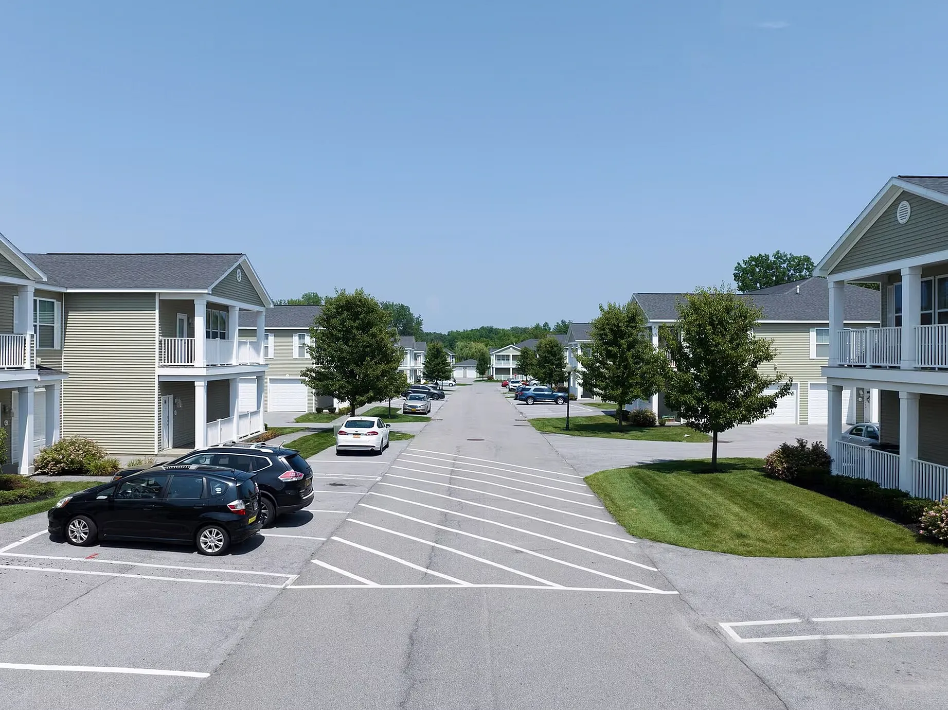 Charming residential street with modern two-story apartments, lush green lawns, and ample parking spaces under a clear blue sky.