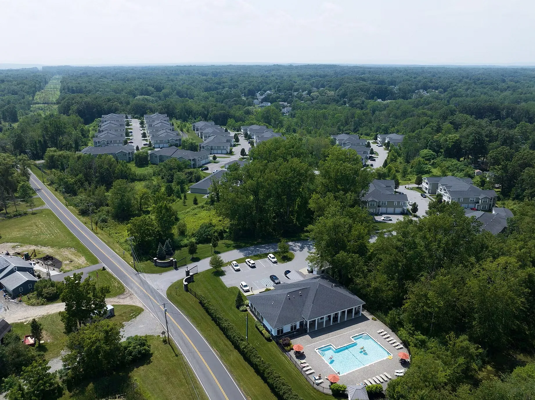 Aerial view of a serene residential community surrounded by lush greenery featuring neatly arranged homes, a spacious clubhouse, and an inviting swimming pool with lounge chairs.