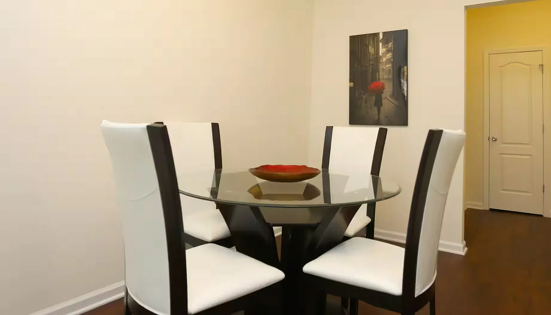 Modern dining area with a round glass table and elegant white chairs, accented by a chic wall painting and warm wooden flooring.