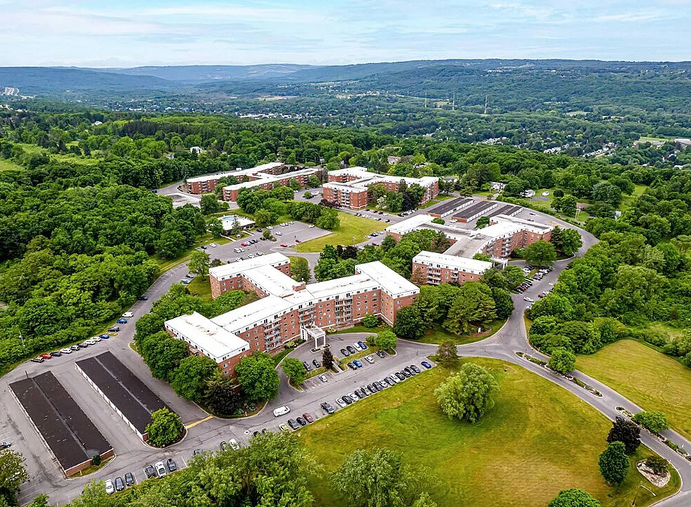 Aerial view of scenic apartment complex nestled in lush greenery and rolling hills, featuring ample parking and sprawling landscaped areas.