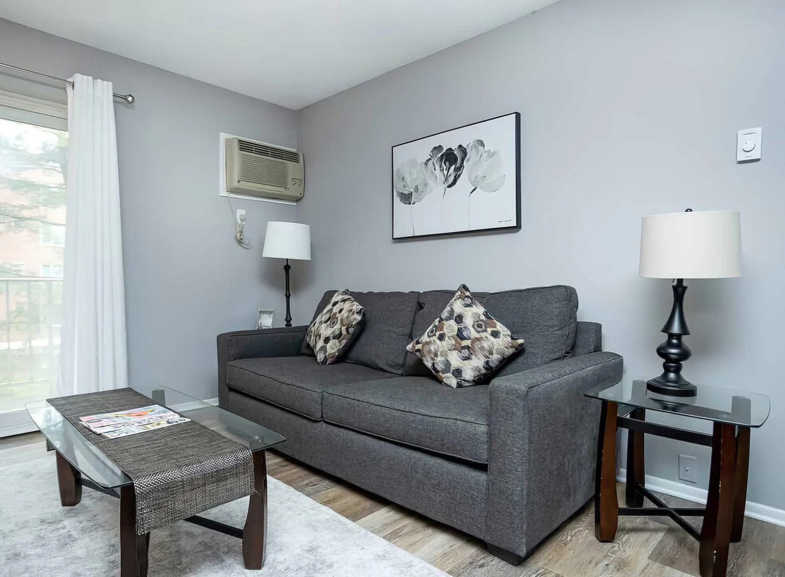 Inviting living room featuring a stylish gray sofa with decorative cushions, modern art, and elegant glass-topped tables, creating a cozy yet contemporary ambiance.