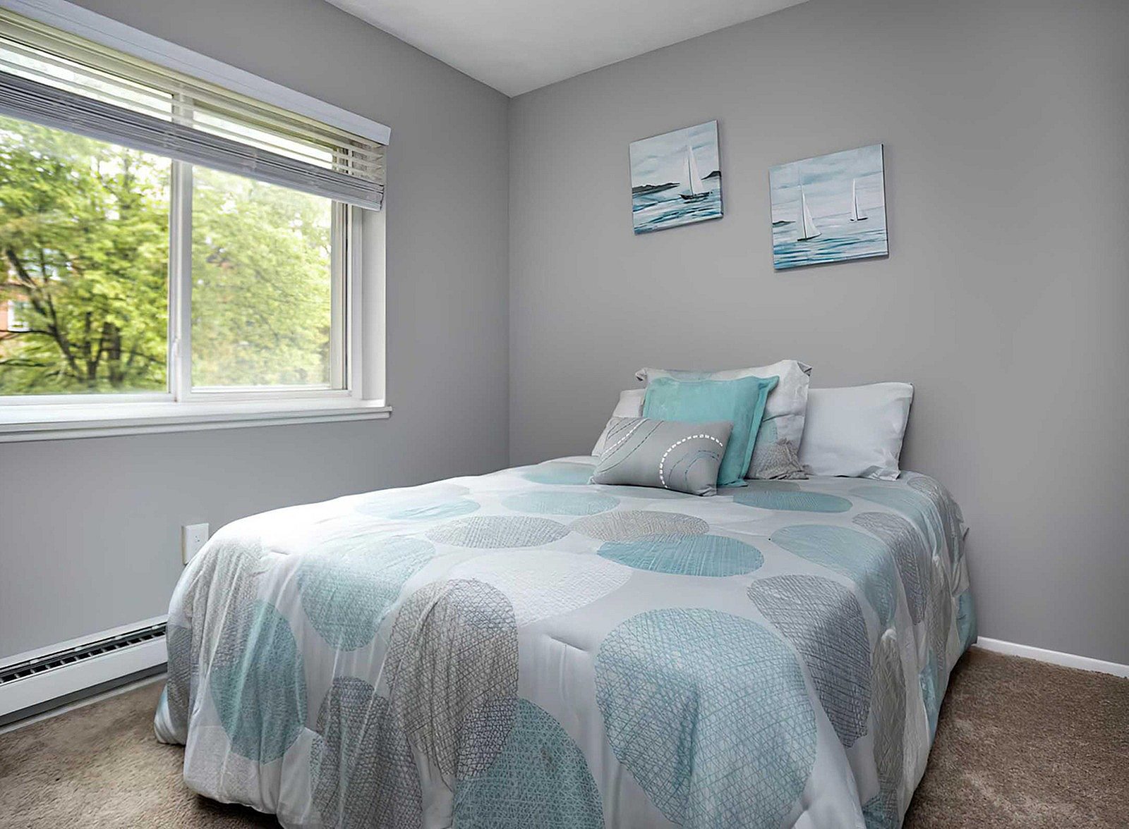 Bright bedroom with modern decor, featuring soft pastel bedding and large window with lush outdoor view.