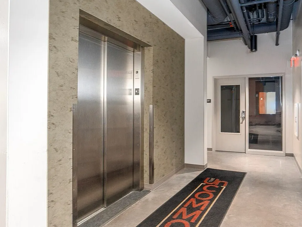 Modern apartment hallway featuring a sleek elevator entrance and high ceilings with industrial-style exposed piping.