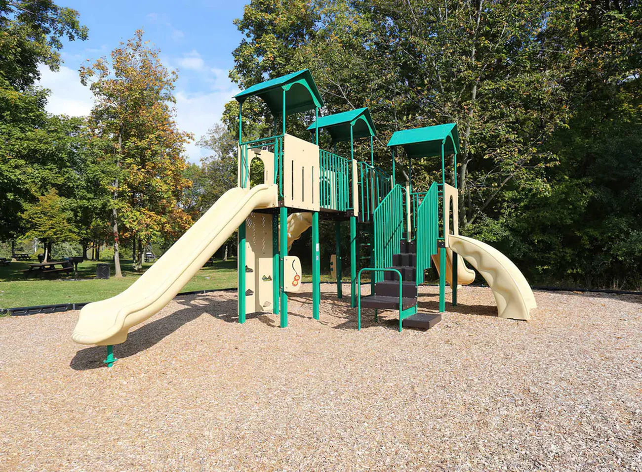 Charming outdoor playground featuring vibrant green and beige structures with slides and climbing areas, surrounded by lush trees.
