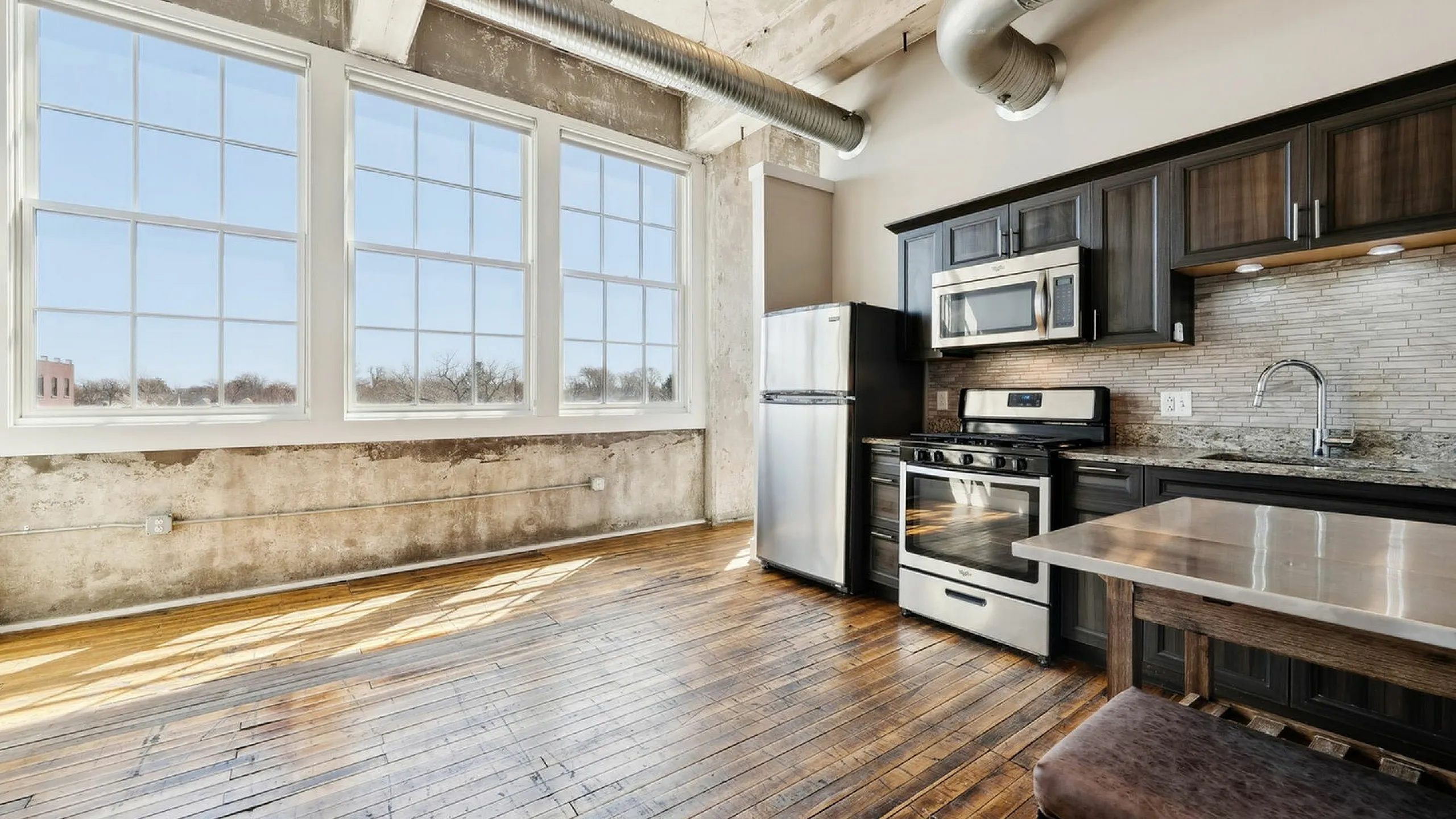 Chic industrial-style kitchen with expansive windows, stainless steel appliances, rustic hardwood floors, and exposed ductwork.