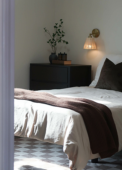 Elegant bedroom with cozy white bed, soft brown blanket, and a warm wall lamp, complemented by a stylish bedside table with decorative greenery.