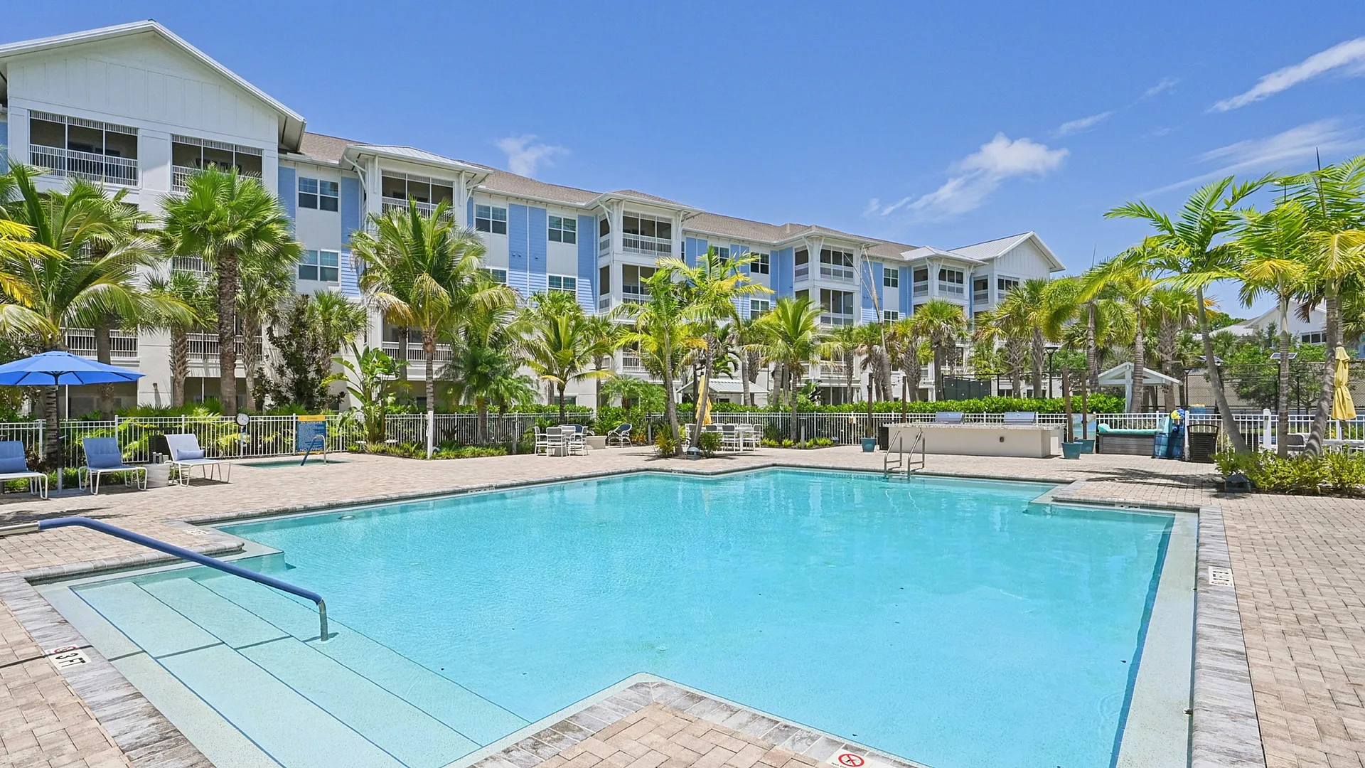 Luxurious apartment complex with a stunning outdoor pool surrounded by lush palm trees, featuring elegant architecture under clear blue skies.