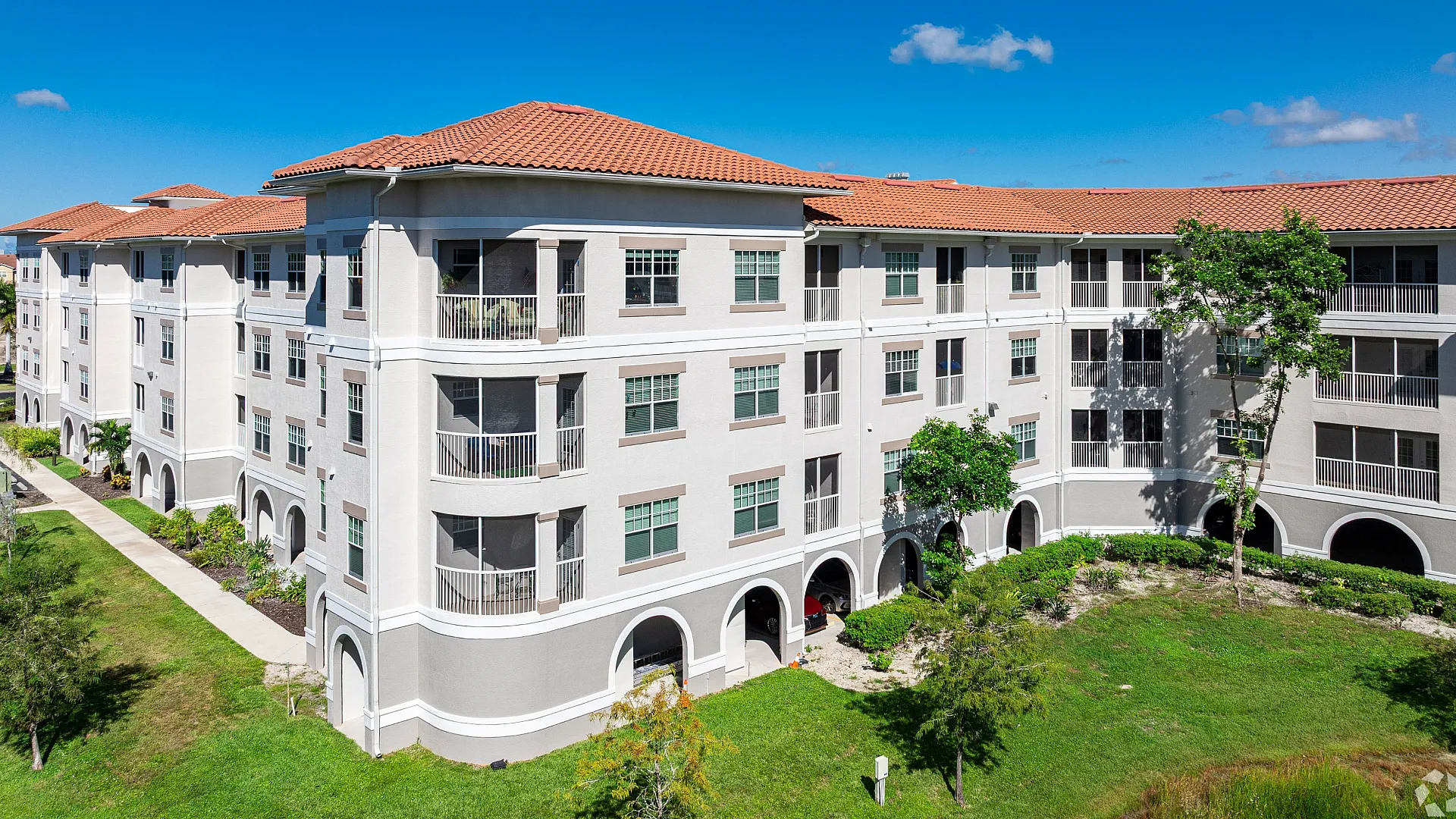Elegant apartment complex with Mediterranean-style architecture featuring red-tiled roofs and lush green surroundings under a bright blue sky.