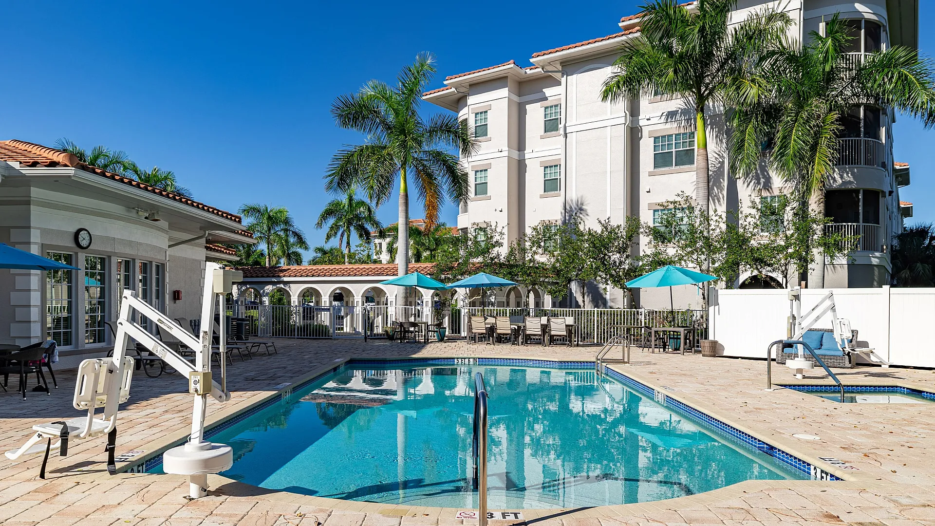 Luxurious apartment complex with inviting swimming pool, surrounded by palm trees and stylish outdoor seating under vibrant blue umbrellas.