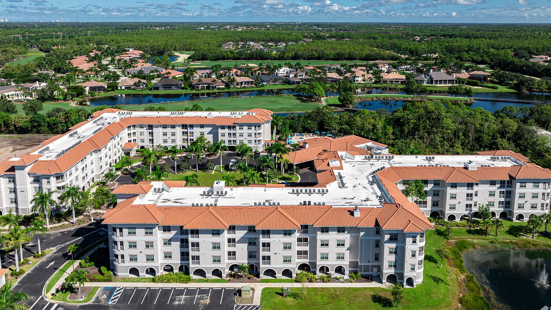 Aerial view of a stunning apartment complex with orange tiled roofs, surrounded by lush greenery and serene water features, offering a tranquil living environment.