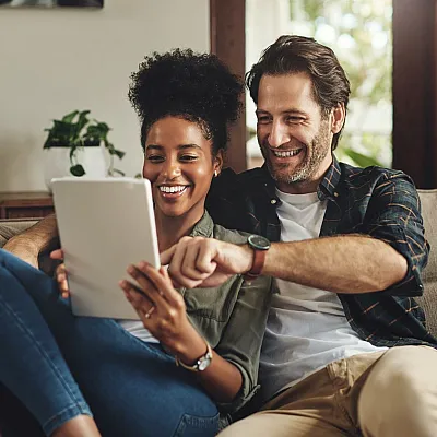 Happy couple enjoying a cozy moment on the sofa, sharing joyful expressions while browsing on a tablet.