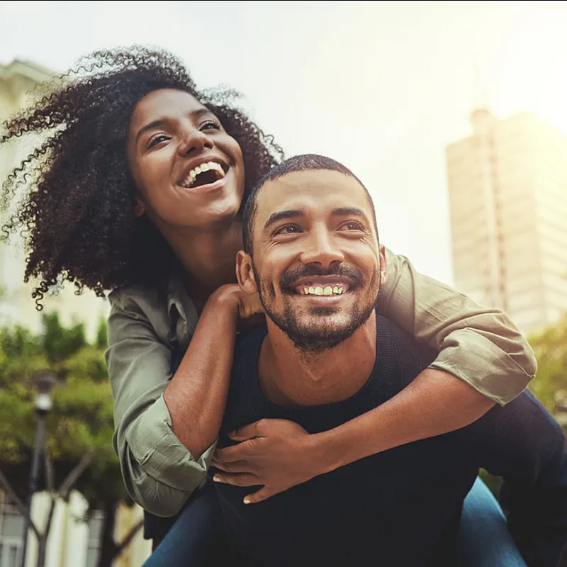 Joyful couple enjoying a playful moment in an urban setting with sunlight highlighting modern architecture in the background.