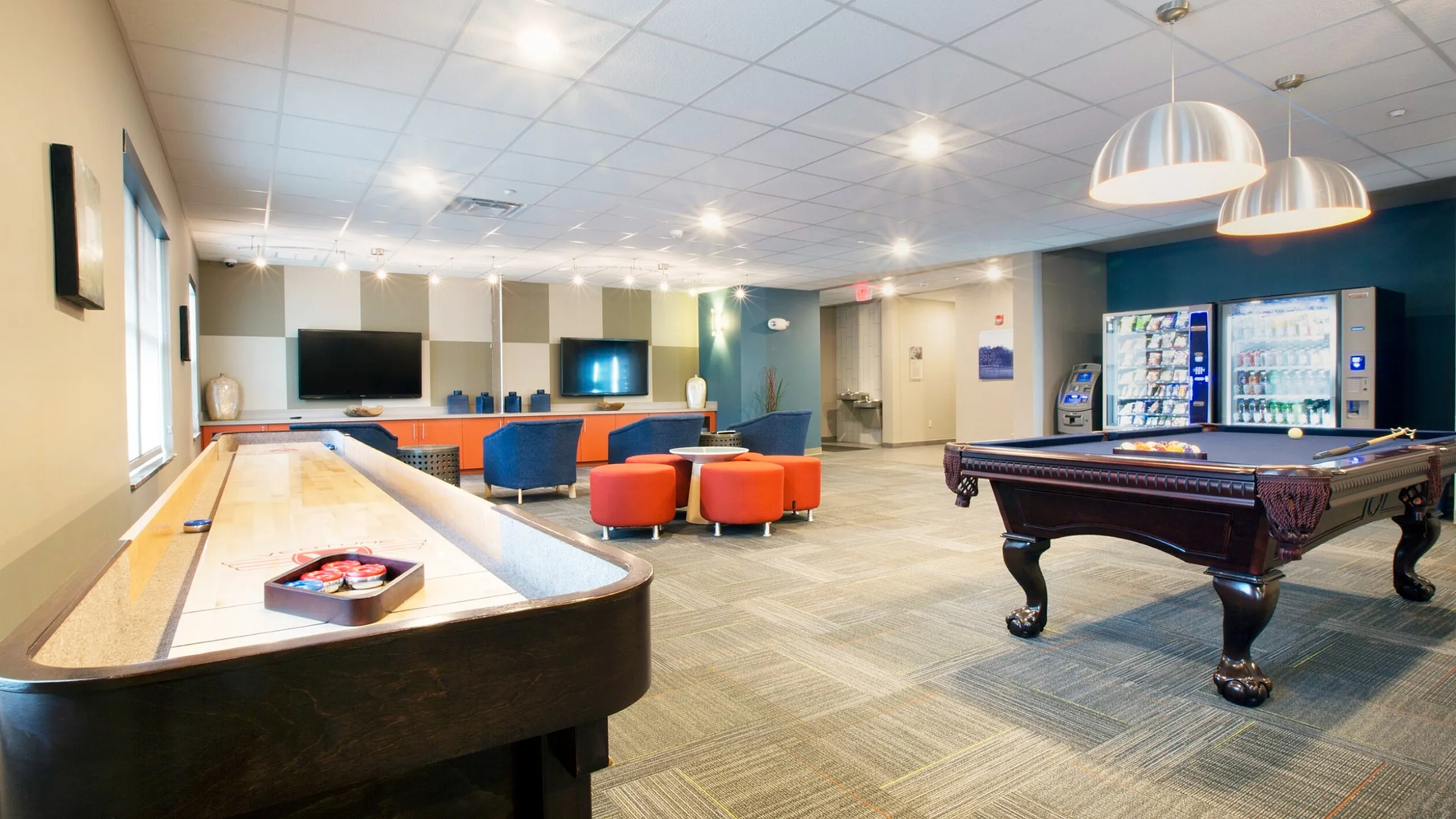 Luxurious apartment recreation room with modern lighting, featuring a pool table, shuffleboard, cozy seating area, and vending machines for ultimate entertainment.