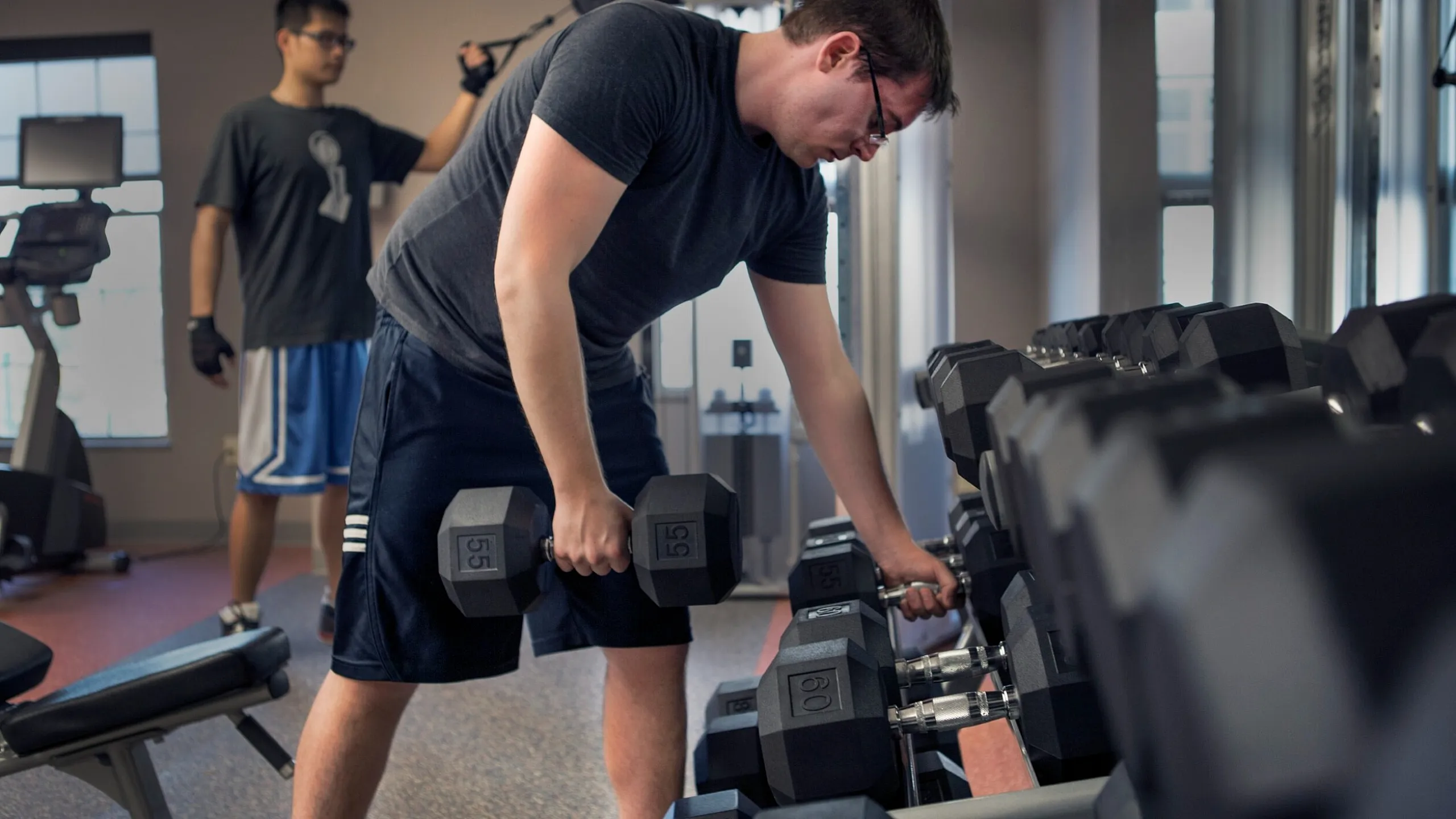 Modern fitness center with a variety of dumbbells and gym equipment, featuring individuals engaged in strength training in a well-lit, motivating environment.