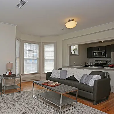 Inviting living room with elegant sofa, stylish coffee table, and bright natural light through large windows, seamlessly connected to a modern kitchen.