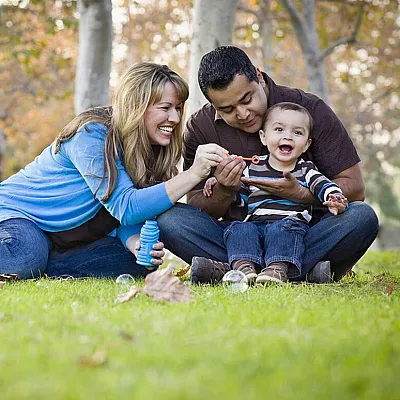 Happy Kensington family enjoying a sunny day in the park, surrounded by lush trees and vibrant green grass, with parents playing with their cheerful child.