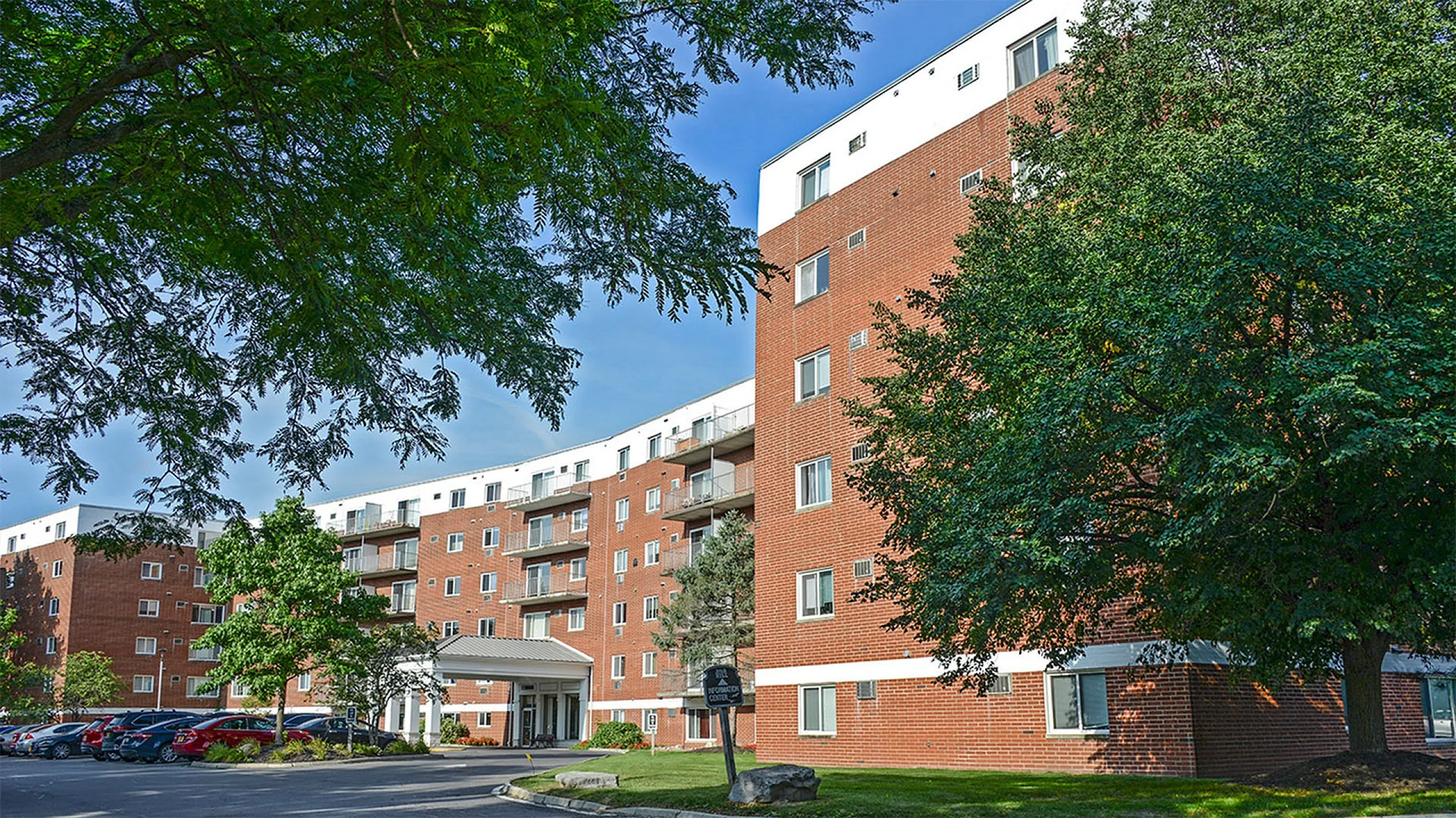 Inviting brick apartment building surrounded by lush green trees, featuring ample parking and welcoming entrance.