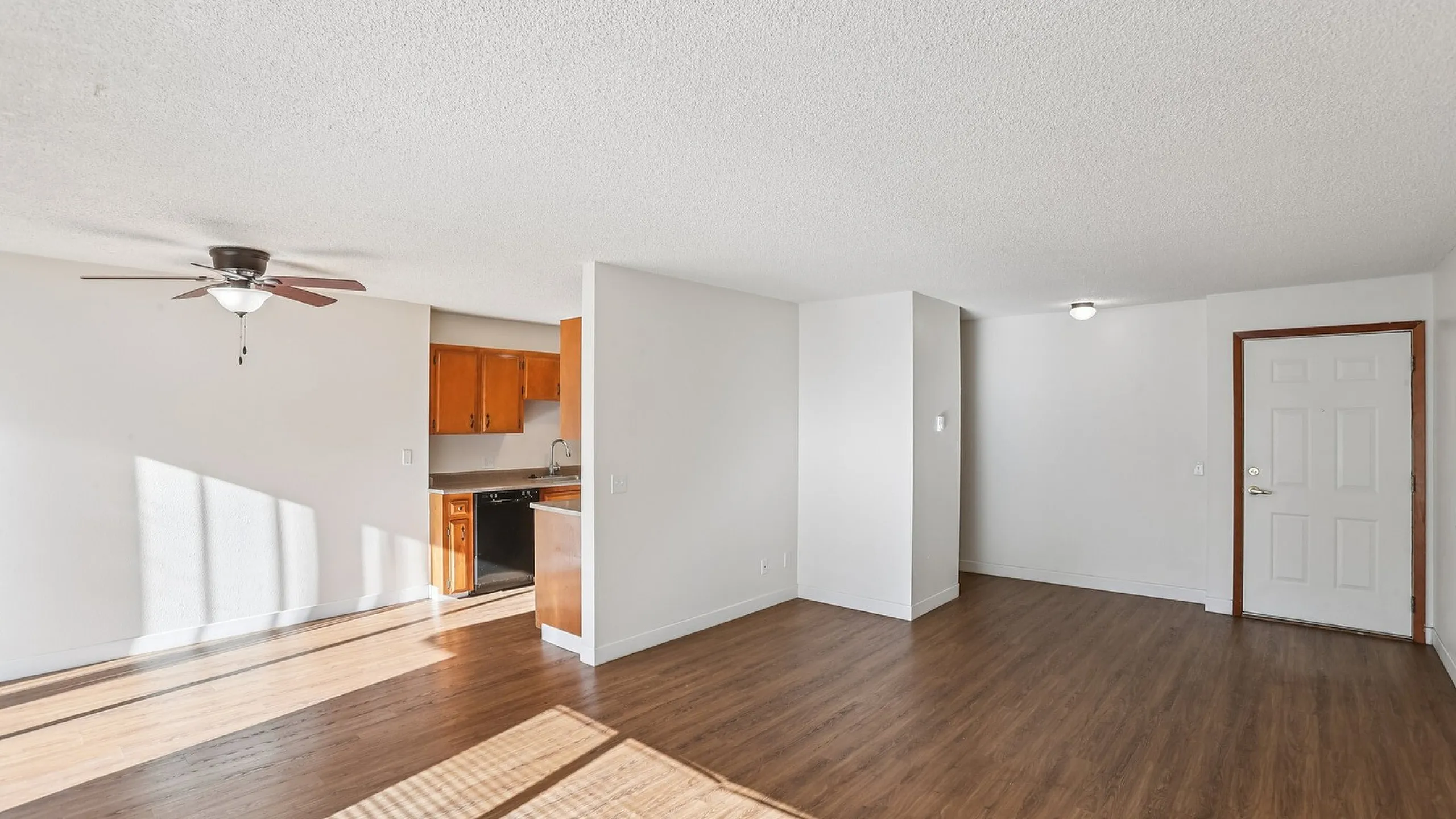 Bright open living area with sleek hardwood floors, modern ceiling fan, and a cozy kitchen featuring elegant wood cabinetry and ample natural light.