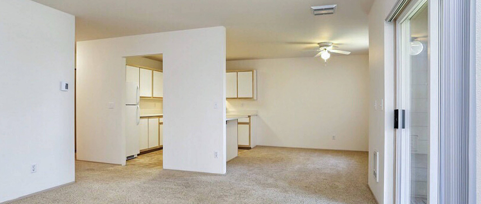 Bright open-concept living area with plush carpet, adjacent to a modern kitchen with ample cabinetry and a ceiling fan for comfort, featuring sliding glass doors for outdoor access.
