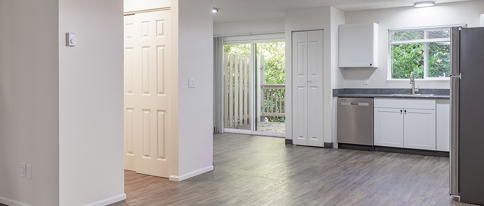 Modern apartment interior with sleek wood flooring, bright open-plan kitchen with stainless steel appliances, and sliding doors leading to a private balcony with lush greenery views.