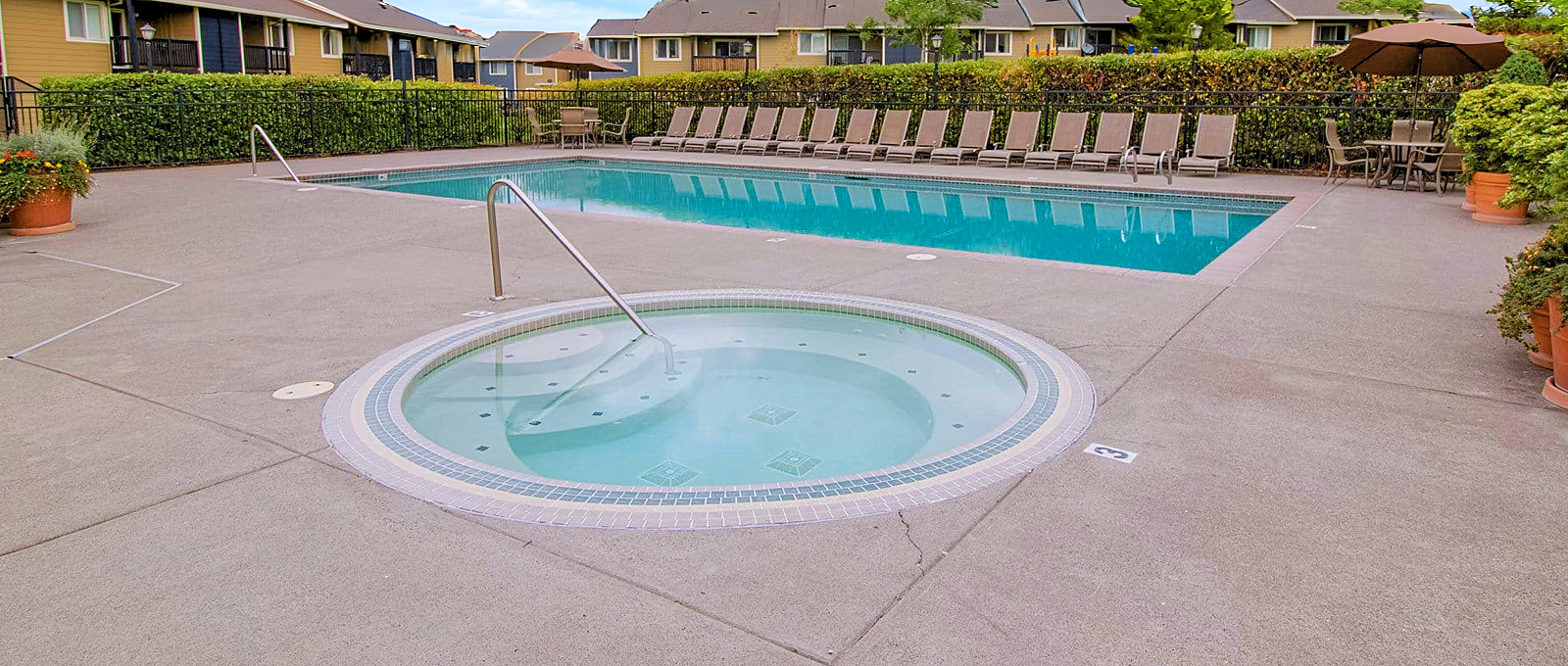 Inviting outdoor pool area with a spacious sun deck, lounge chairs, and a hot tub, perfect for relaxation in a serene apartment community.