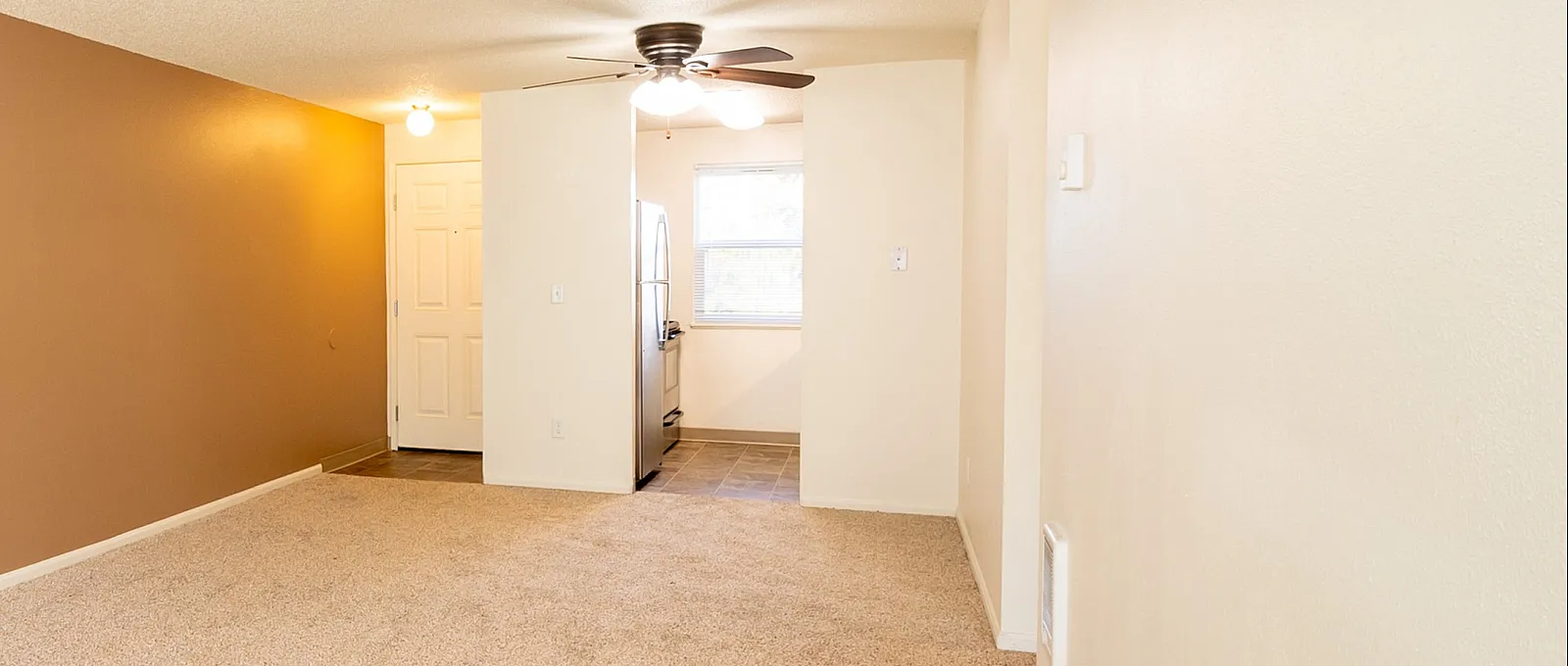 Spacious living area with warm beige carpet and stylish accent wall, leading to a modern kitchen illuminated by natural light.
